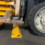 Thumbnail of http://A%20closeup%20of%20a%20of%20a%20Forklift%20Wheel%20Stop%20Mounted%20on%20the%20Ground%20of%20a%20Warehouse%20in%20Front%20of%20a%20Pallet%20Wrapper%20Machine%20With%20a%20Forklift%20Lifting%20a%20Pallet%20over%20the%20Stop